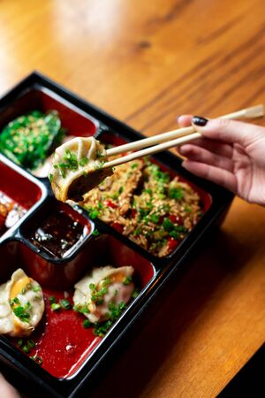 Set of chinese asian dishes on one plate standing on a wooden tabletop. A woman's hand holds a fried dumpling with a chopsticks over a dish. Vertical frame.の写真素材