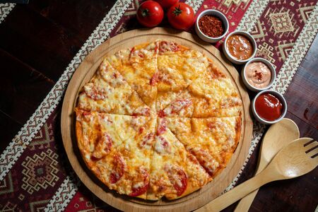 Pizza lying on a wooden plate on a tablecloth with national patterns, next are four sauces and three tomatoes.  Wooden fork and spoon lying around.の写真素材