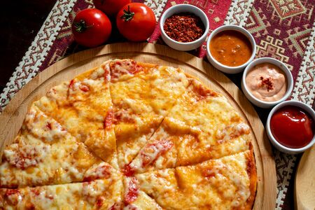 Pizza lying on a wooden plate on a tablecloth with national patterns, next are four sauces and three tomatoes.の写真素材