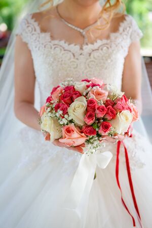 The bride in a white wedding dress with a red ribbon on her belt, with a wedding bouquet of roses in her hands.の写真素材