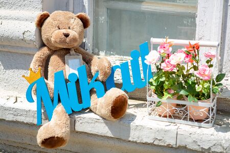 Brown Teddy bear sitting on the windowsill with a blue sign in his paws, next to two pots of flowers.の写真素材