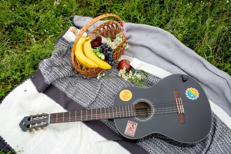 Black acoustic guitar lying on a gray-white blanket next to a wicker basket of fruit and a bottle of wine.の写真素材