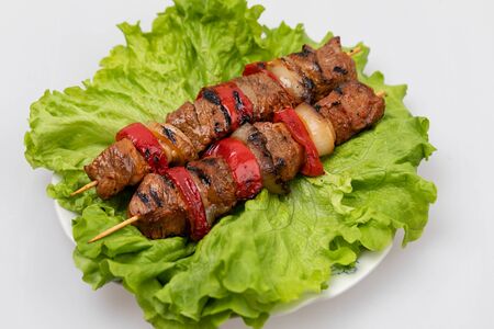 Pieces of fried meat and vegetables on skewers lying on a plate on a sheet of salad on a white background.の写真素材