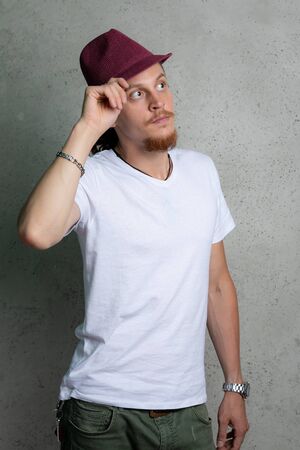 A guy in a white t shirt with a Burgundy hat, thinking, on a gray background.の写真素材