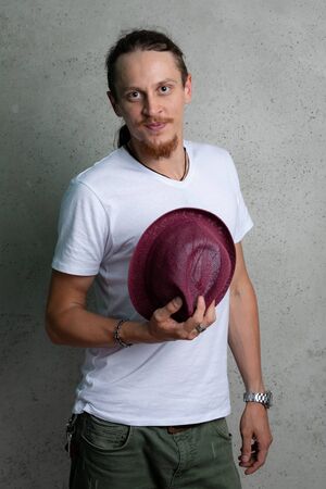 A guy in a white tshirt with a Burgundy hat in his hand, on a gray background.の写真素材