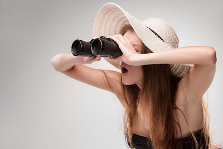Young woman in hat looking through binoculars on gray background. Travel and adventure concept. Closeup.の写真素材