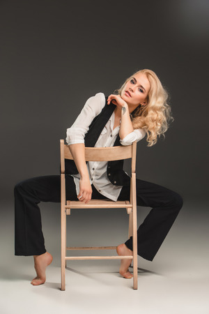 Beauty blond woman  in a black suit and white shirt, sitting on a chair on a gray backgroundの写真素材