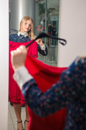Woman trying red dress shopping for clothing. Beautiful happy blonde smiling young shopper woman looking in mirror standing in clothes store.の写真素材