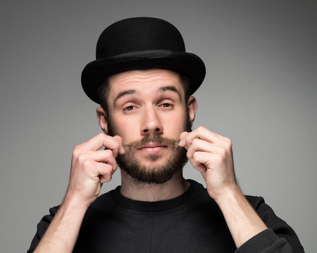 Young attractive macho stylish fashionable guy.  portrait on gray background. a man in a black hat with a beard mustache curls. concept of narcissismの写真素材
