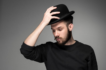 Young man with a mustache and beard raising his hat  in respect and admiration for someone. portrait on gray background. male gaze directed downwardの写真素材
