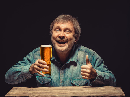 Enjoying his favorite beer.  The front view of handsome smiling  man as fan in denim shirt with glass of beer, sitting at the wooden tableの写真素材