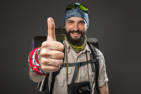 Portrait of a male smiling fully equipped tourist with backpack and the camera on gray background. the focus arm with a gesture of approvalの写真素材