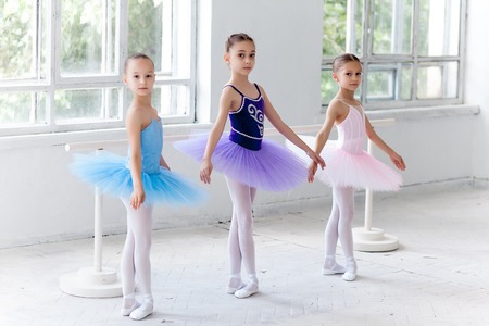 Three little ballet girls in multicolored tutu posing at ballet barre together on white backgroundの写真素材