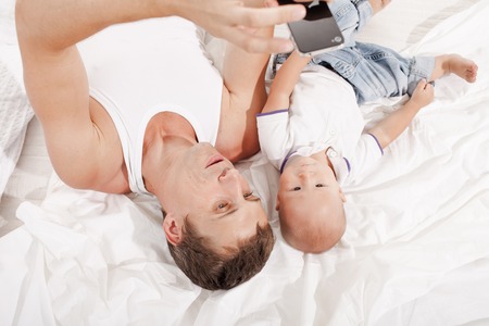 Young smiling father with his nine months old son  lying upside down on the bed at home on white home backgroundの写真素材
