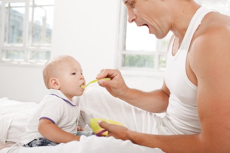 Young smiling father with his nine months old son on the bed at home on white home background. Dad feeding his son with a spoonの写真素材