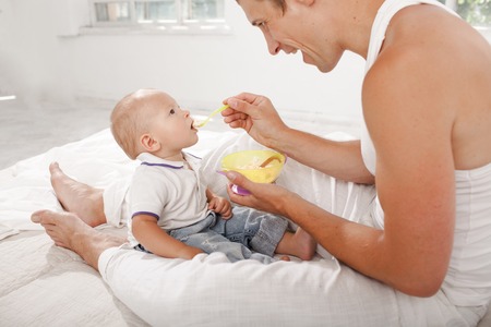 Young smiling father with his nine months old son on the bed at home on white home background. Dad feeding his son with a spoonの写真素材