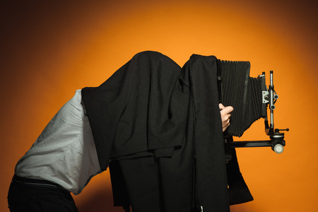 photographer hiding under a black cape with retro camera on an orange backgroundの写真素材