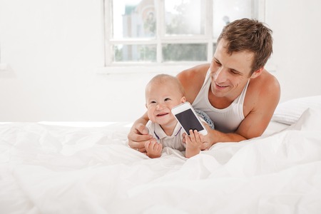 Young smiling father with his nine months old son on the bed at home on white home background