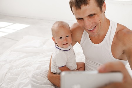 Young smiling father with his nine months old son on the bed at home on white home background. dad taking selfie photo with telephone cameraの写真素材