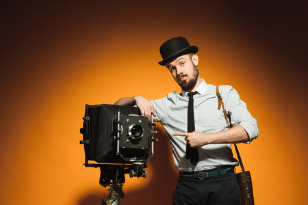 Young  positive man in hat as photographer with retro camera on an orange backgroundの写真素材