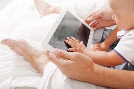 Young father with his nine months old son sitting on the bed at home on white home background with digital tabletの写真素材