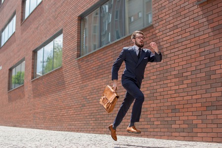 Young businessman with a briefcase and glasses running in a city street on a background of red brick wallの写真素材