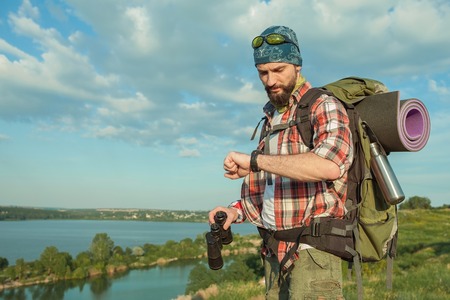 Young caucasian tourist with backpack  standing on the top of hill and looking at  watch. The concept of an invitation to travelの写真素材