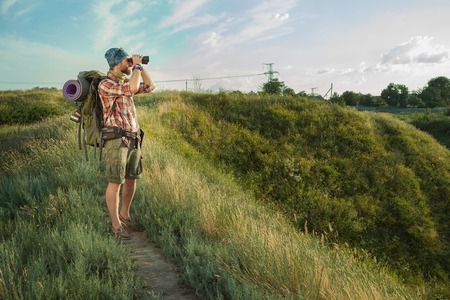 Young smilimg caucasian tourist with backpack  standing on the top of hill against blue sky background and looking through binocularsの写真素材