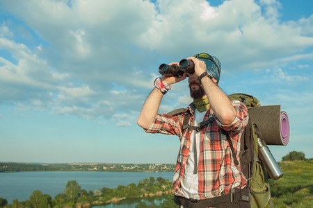 Young smilimg caucasian tourist with backpack  standing on the top of hill against blue sky background and looking through binocularsの写真素材