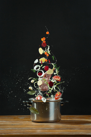 Fresh vegetables falling into a stainless steel casserole on wooden table. The concept of cooking borsch