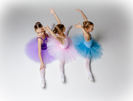 Three little ballet girls sitting in multicolored tutus and pointe shoes together on white backgroundの写真素材