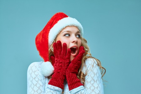 The surprised and happy girl dressed in santa hat. Holiday concept with blue background.の写真素材