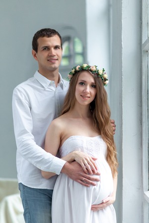 Cheerful young couple dressed in white standing against white home. Husband hugging his pregnant wifeの写真素材
