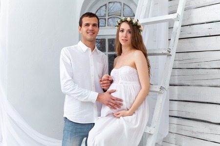 Cheerful young couple dressed in white standing against white home. Husband hugging his pregnant wifeの写真素材