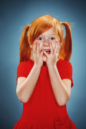 The beautiful portrait of a  surprised little girl with red hair in red dress on blueの写真素材