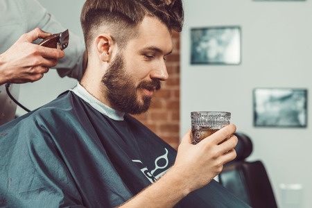 The hands of young barber making haircut of attractive bearded man in barbershopの写真素材