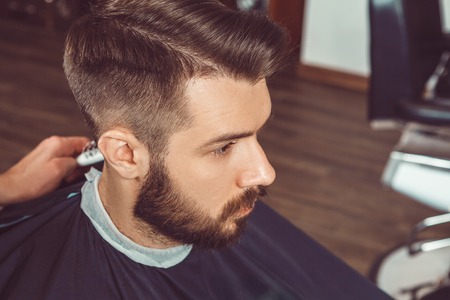 The hands of young barber making haircut of attractive bearded man in barbershopの写真素材