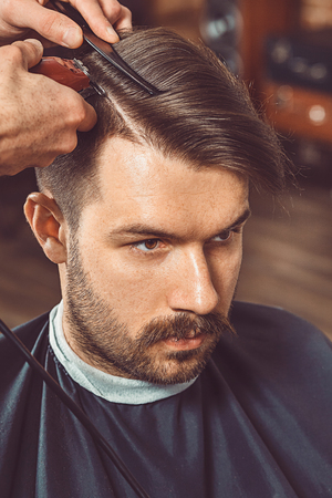 The hands of young barber making haircut of attractive bearded man in barbershopの写真素材