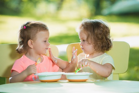 Two little 2 years old girls sitting at a table and eating together against a green lawnの写真素材