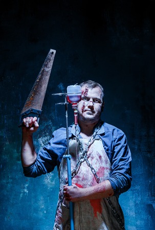 Bloody Halloween theme: crazy killer as bloody butcher with saw on dark blue backgroundの写真素材