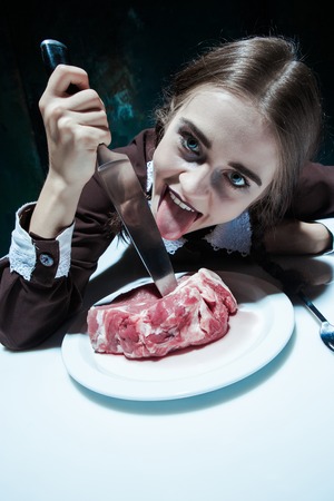 Portrait of a young girl with knife and and a piece of raw meat on a plate. Girl in school uniform as killer. The image in the style of Halloween and Addams familyの写真素材