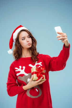 Girl dressed in santa hat with a Christmas gift and phone. She doing selfie picture. Holiday concept with blue background.の写真素材