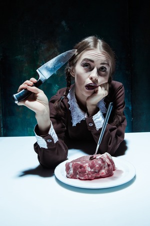 Portrait of a young girl with knife, fork and and a piece of raw meat on a plate. Girl in school uniform as killer. The image in the style of Halloween and Addams familyの写真素材