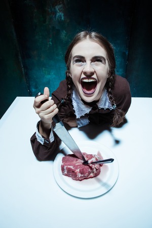 Portrait of a young girl with knife, fork and and a piece of raw meat on a plate. Girl in school uniform as killer. The image in the style of Halloween and Addams familyの写真素材