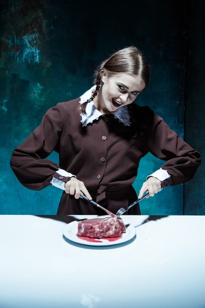Portrait of a young girl with knife, fork and and a piece of raw meat on a plate. Girl in school uniform as killer. The image in the style of Halloween and Addams familyの写真素材