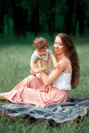Young beautiful mother sitting with her little son against green grass. Happy woman with her baby boy on a summer day. Family walking on the meadow.の写真素材