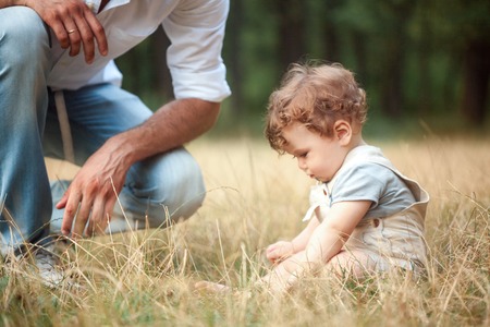 Young beautiful father and little toddler son against green grass. Happy father with her baby boy on a summer sunny day. Family walking on the meadow.の写真素材