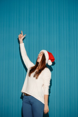 The surprised christmas girl wearing a santa hat pointing up on the blue studio backgroundの写真素材