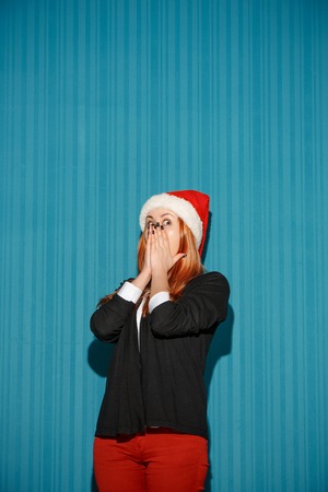 The surprised christmas girl wearing a santa hat on the blue studio backgroundの写真素材
