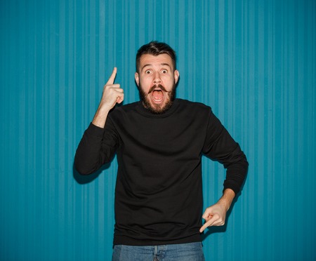 Portrait of young man with shocked facial expression pointing up over blue studio backgroundの写真素材